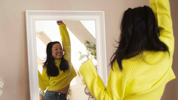 Une femme au pull jaune leve le bras devant le miroir, euphorique.