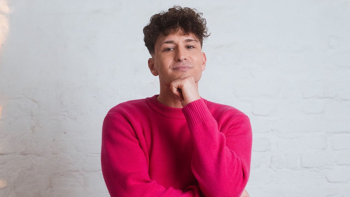 un homme vetu d'un pull rose regarde pensivement la caméra