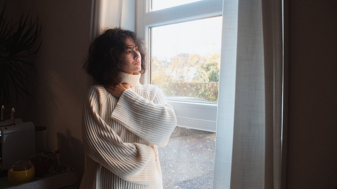 Femme au pull blanc debout devant une fenetre regardant pensivement vers l exterieur.