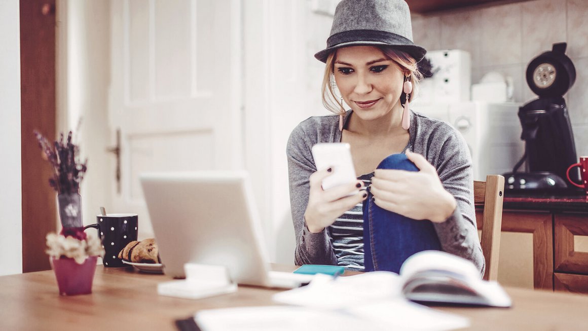 Junge Frau am Tisch mit Smartphone, Notebook und Unterlagen zum Haushaltsbuch führen